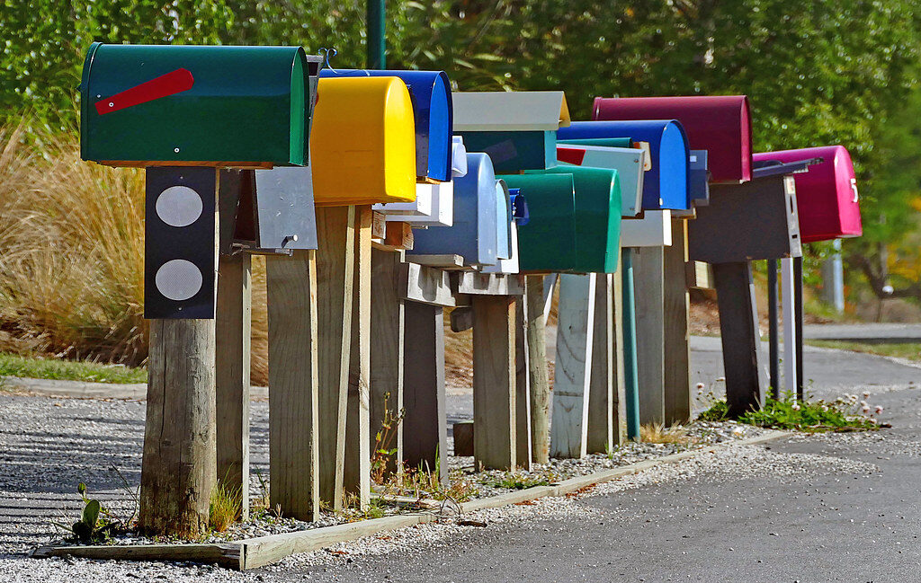 Eine Reihe roter Briefkästen an einer Landstraße, dahinter Felder und Bäume